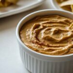 A creamy, smoky chipotle queso dip in a white bowl, garnished with flecks of chipotle peppers and served with crispy tortilla chips.