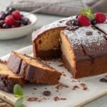 A rich chocolate pound cake dusted with powdered sugar, topped with fresh berries, and served on a wooden board with a bowl of fruit.