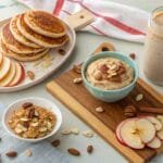A breakfast spread featuring apple slices, pancakes, a smoothie, and bowls of porridge topped with nuts, presented on a light-colored surface.