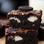 A stack of gooey dairy-free Oreo brownies on a wooden plate, topped with crushed Oreo pieces.